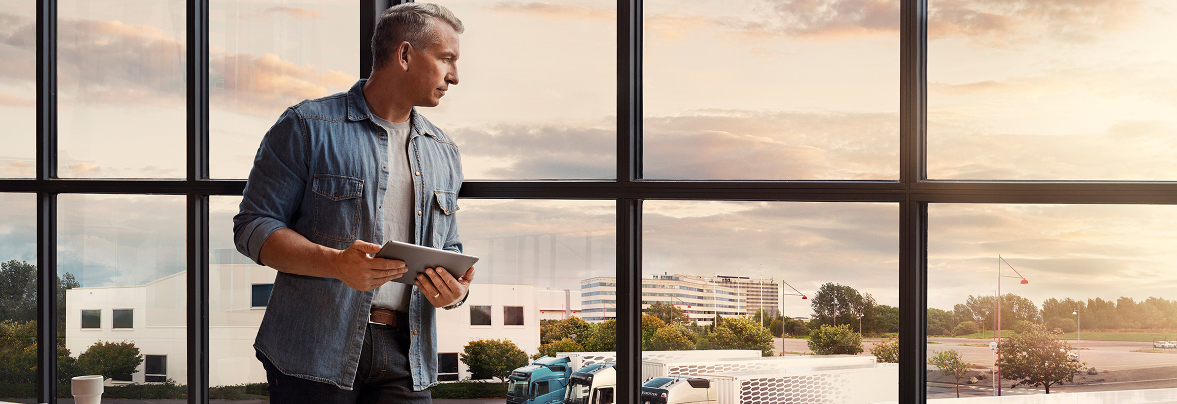 A man holding a tablet stands by a window and looks down over his truck fleet